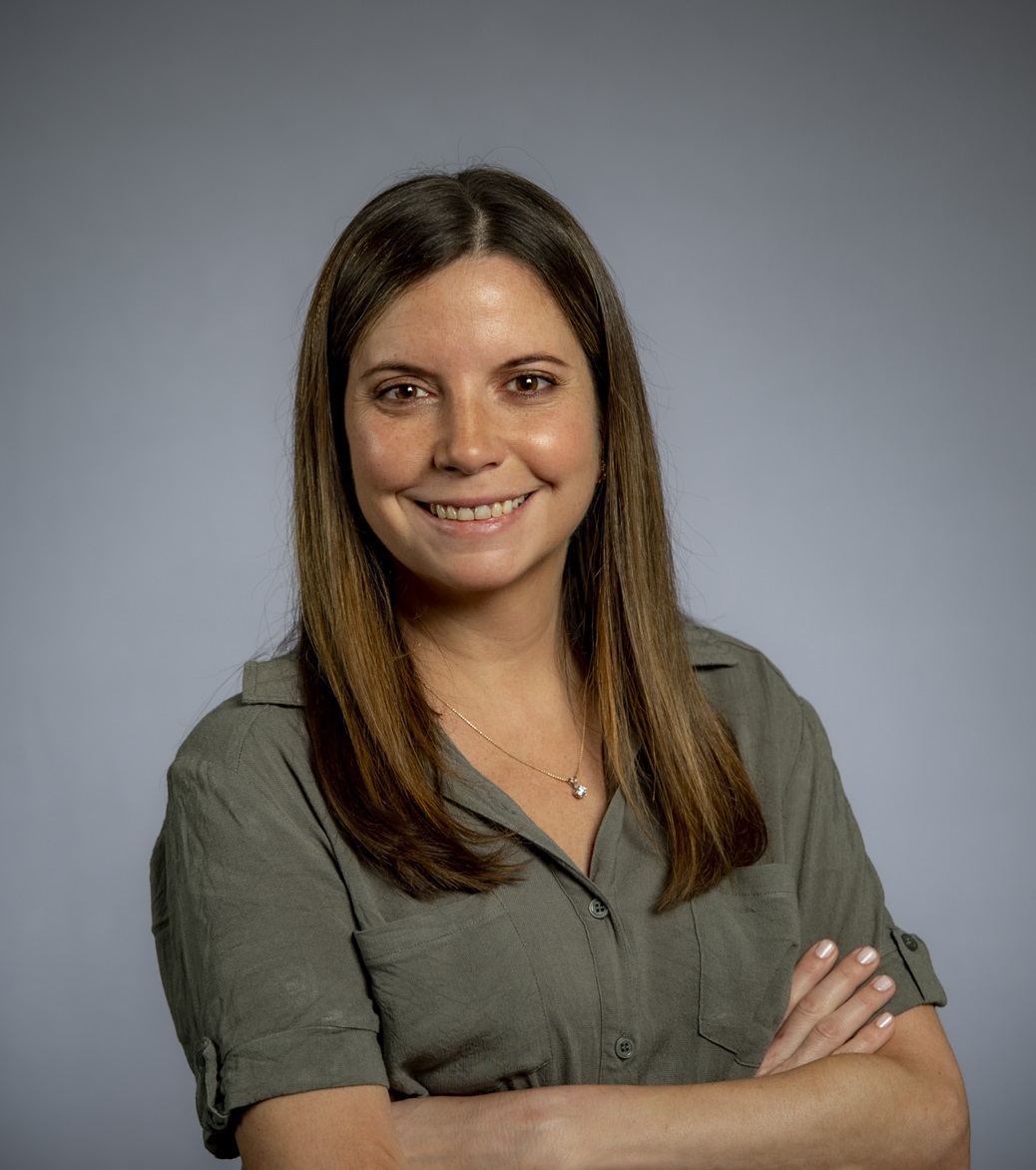 headshot of a smiling woman with long brown hair wearing a green top and crossing her arms against a gray background
